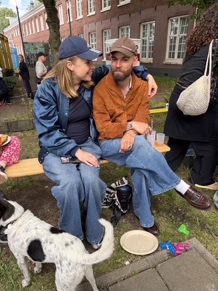 A couple is seen relaxing on a wooden bench outdoors, accompanied by a dog, amidst a casual gathering in the Bruno van Moerkerkenpassage, Rapenburg, Amsterdam
